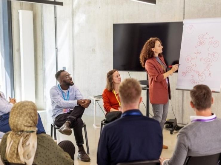 A group of peers discuss something with a teacher, while the teacher gestures at a whiteboard.
