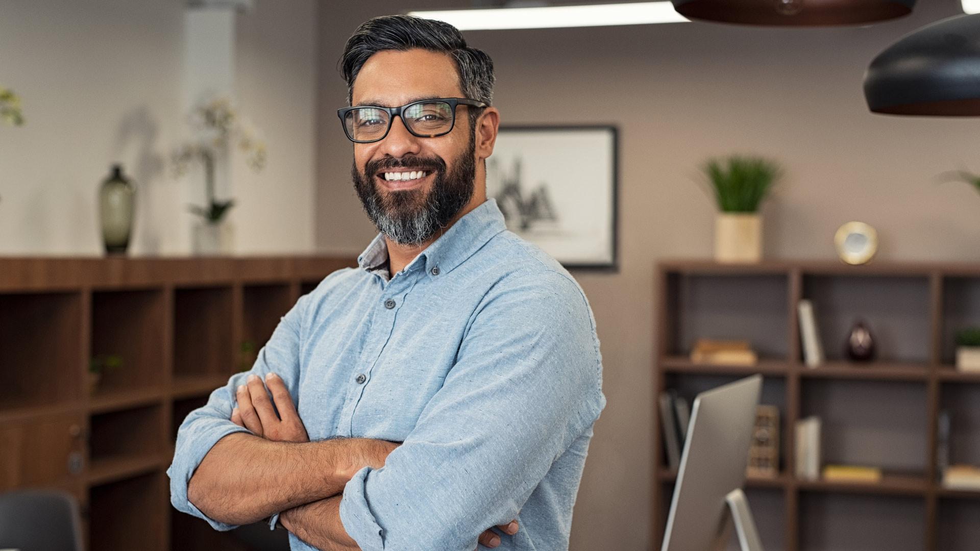 Confident civil engineering professional in a modern office, wearing a light blue button-up shirt and glasses, smiling with arms crossed—representing leadership and innovation in engineering.