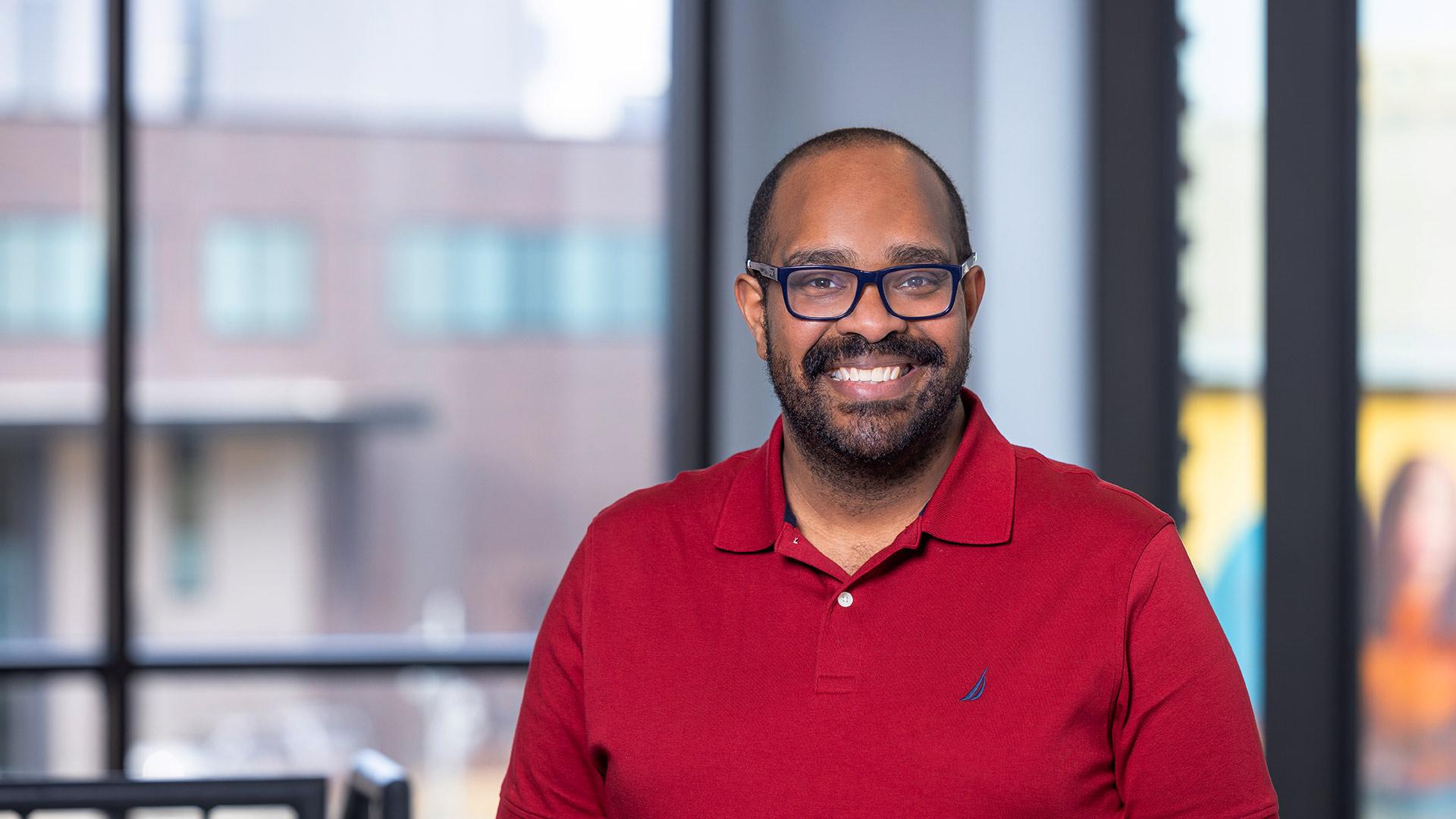 Smiling professional in a red polo shirt standing in a modern office with large windows—representing leadership, education, and career growth in the MA in Leadership for Educational Organizations program.