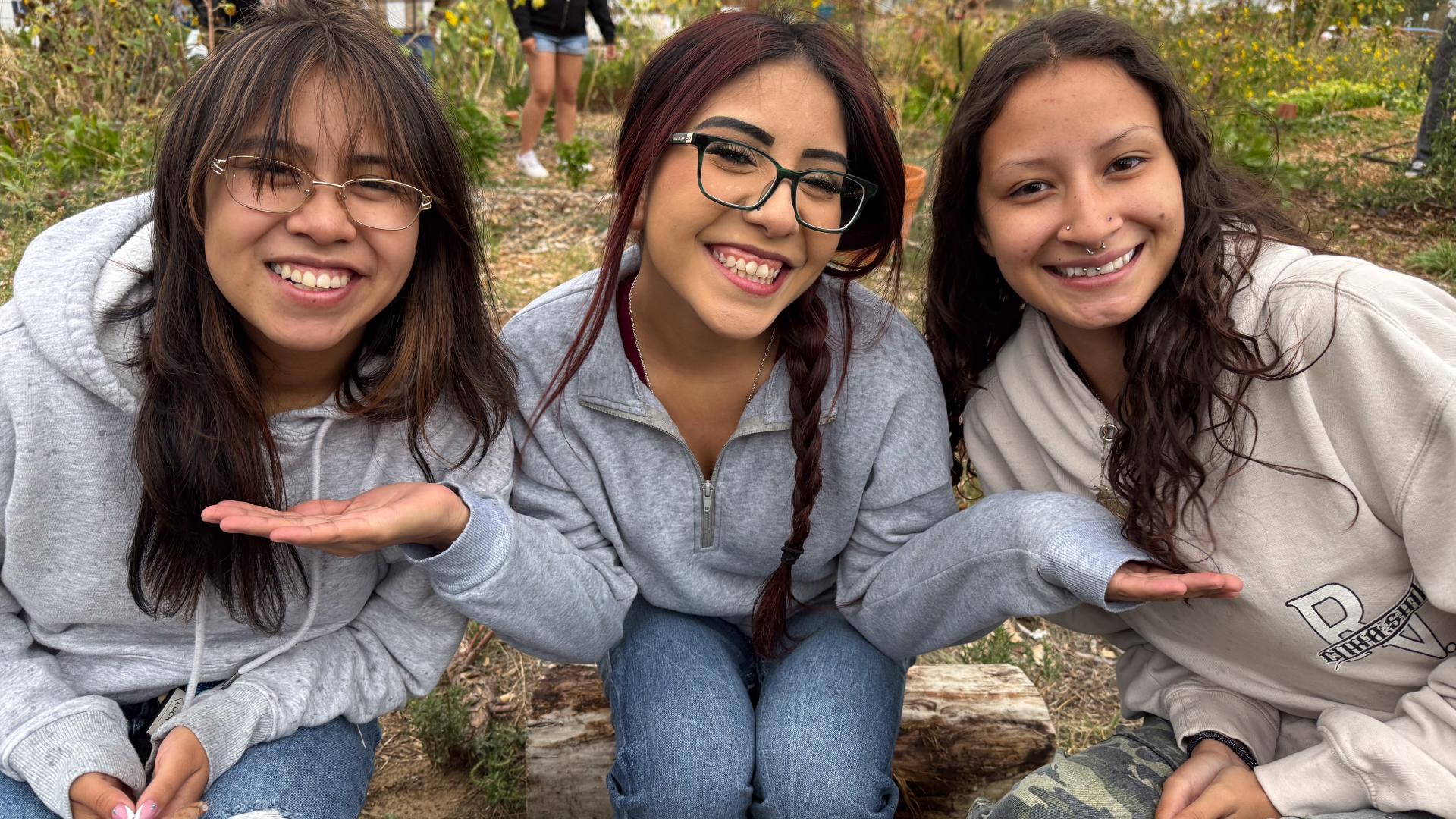 3 female anthropology students sit together smiling with green foliage in the background.