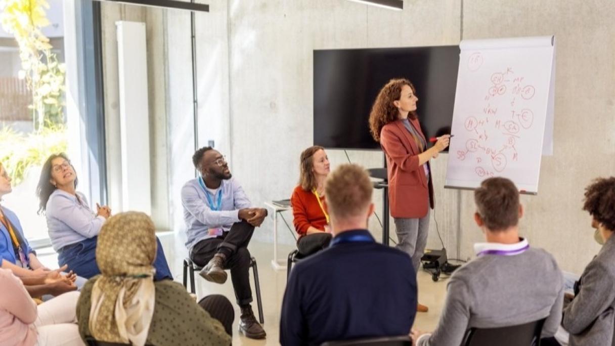A group of peers discuss something with a teacher, while the teacher gestures at a whiteboard.
